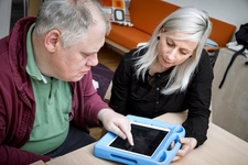 Foto: Mann bedient Kommunikations App am iPad und LIFEtool Beraterin Carina Bloder begleitet ihn dabei.